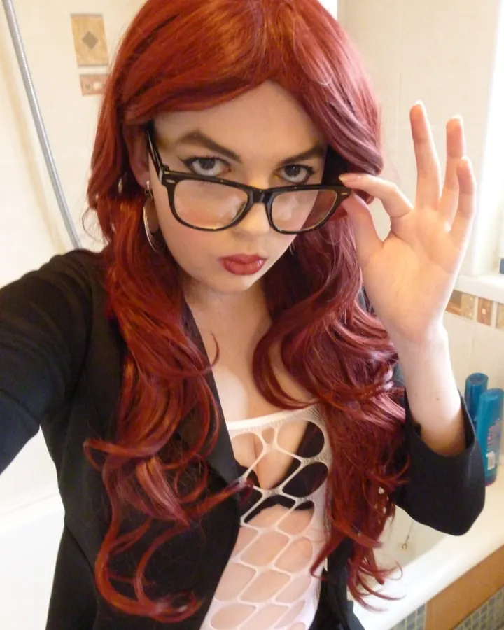 Femme aux cheveux bruns, vêtue d'une tenue noire, posant devant un rideau blanc, mains sur les hanches, regard direct.