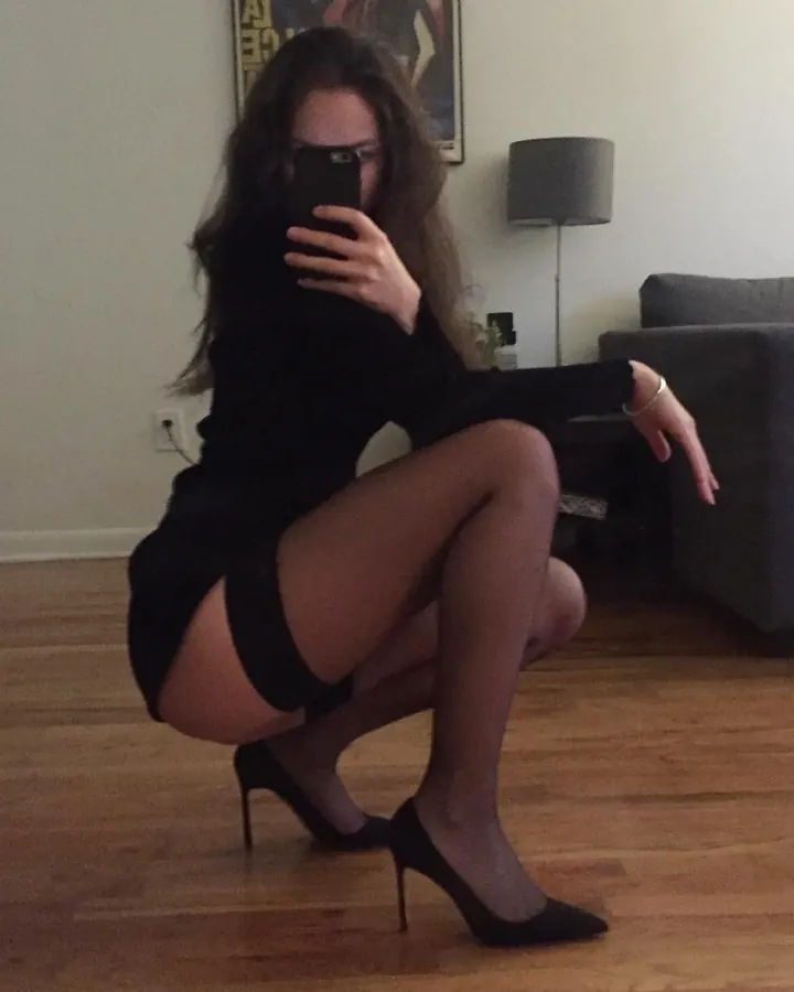 Femme aux cheveux bruns, vêtue d'une tenue noire, posant devant un mur, mains sur les hanches, regard direct.