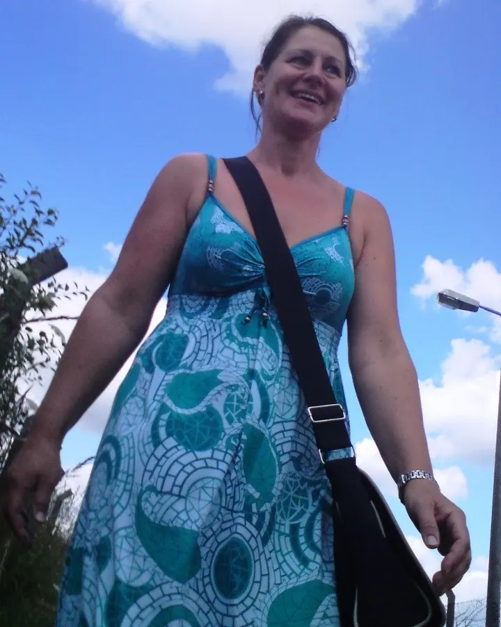 Femme mûre aux cheveux châtains et yeux bleus, souriant doucement sous un ciel bleu, en robe turquoise à motifs géométriques.