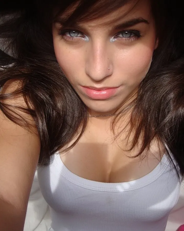 Femme aux cheveux bruns foncés et yeux bleus, en haut blanc, regardant l'appareil de près, avec un piercing au nez.