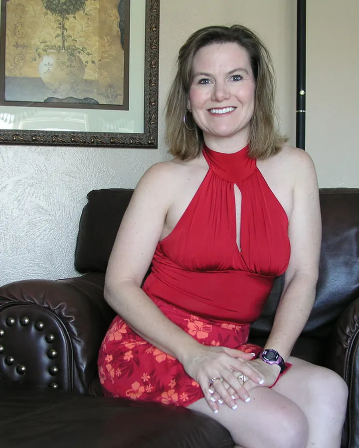 Femme aux cheveux châtains et yeux bleus, en robe rose vif, assise sur un canapé ancien, souriante, en sandales noires.