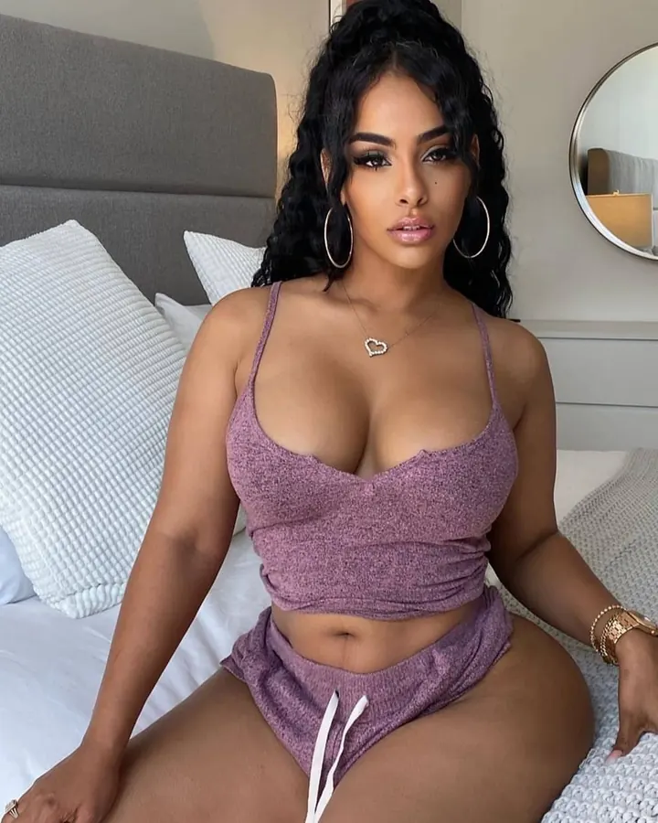 Femme ronde en lingerie noire, posant sur un lit avec une main sur la poitrine, sous une lumière naturelle.