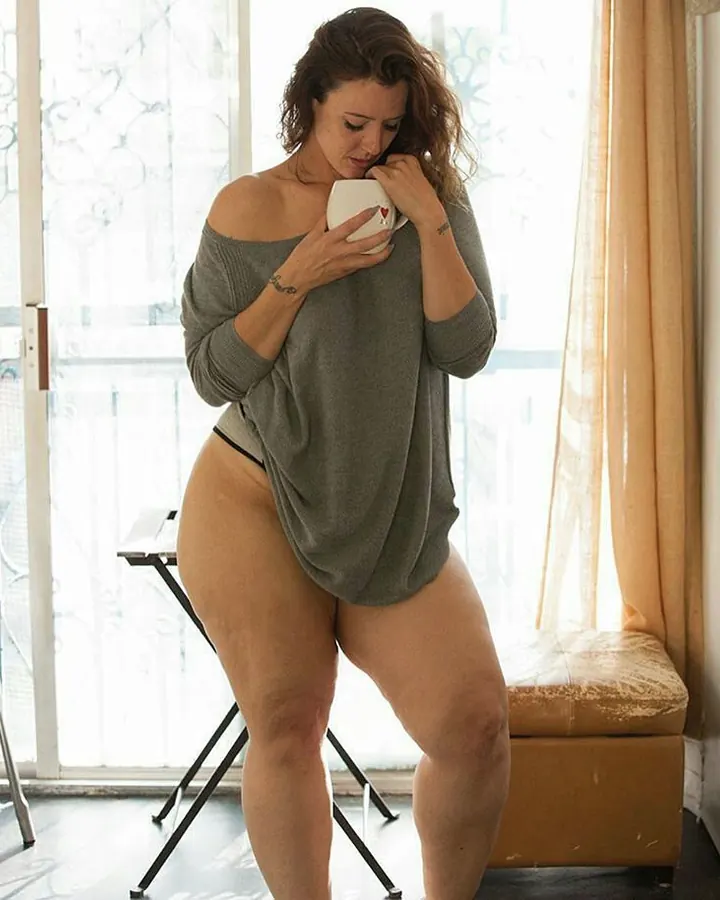 Femme ronde nue, assise sur un canapé gris, tenant une branche de fleurs blanches, avec une fleur dans les cheveux.