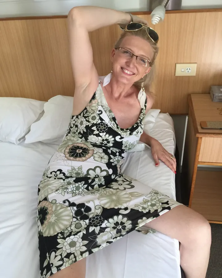 Femme aux cheveux blonds, vêtue d’une robe à motifs floraux, assise sur un lit, les lunettes de soleil sur la tête, souriant à la caméra.