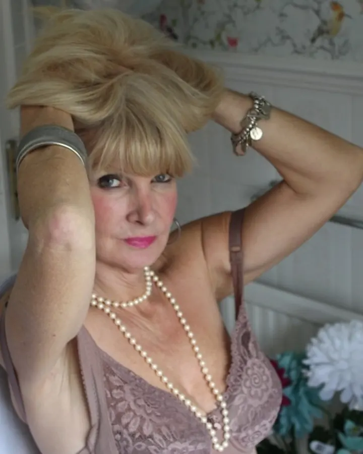 Femme aux cheveux blonds courts, vêtue d’un haut noir sans manches, assise sur un canapé fleuri, les jambes croisées, portant des sandales à talons.