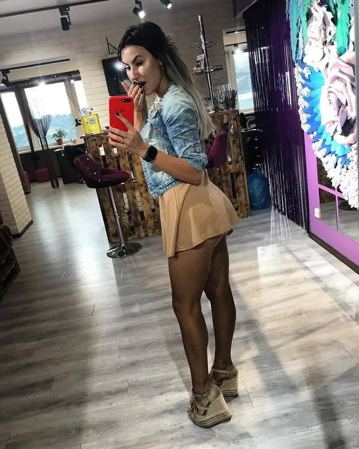 Femme aux cheveux bruns foncés, vêtue d’un haut bleu clair et d’une jupe beige, tenant un téléphone rouge, debout dans une pièce lumineuse avec des meubles en bois.