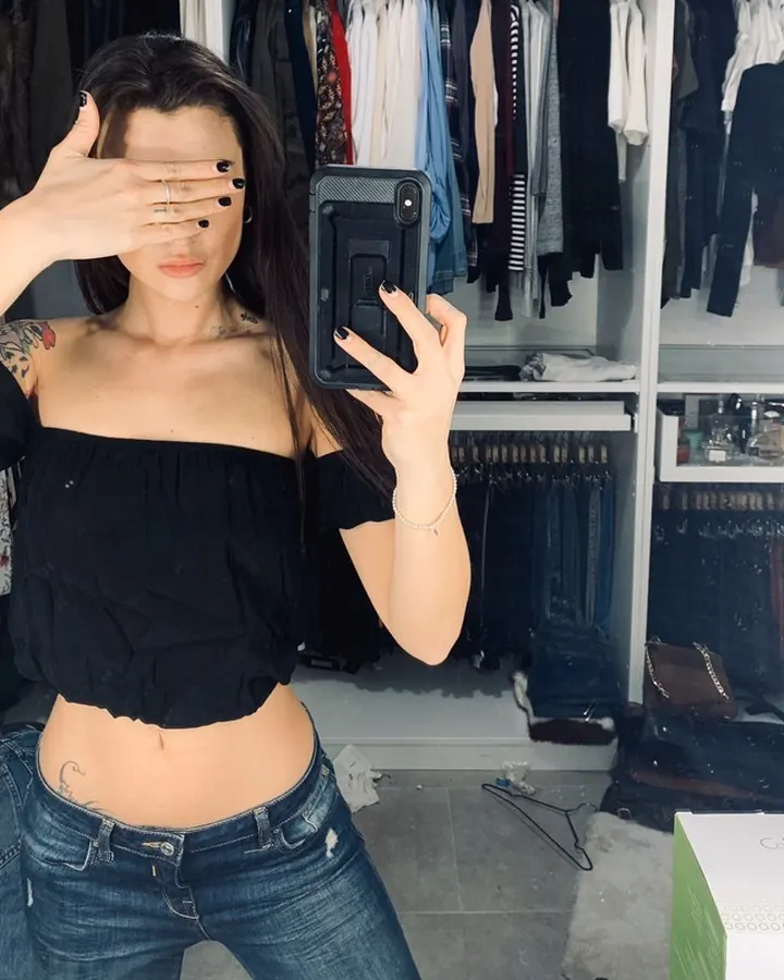 Femme aux cheveux châtains, portant un haut noir et un short en jean, prenant un selfie dans un dressing.