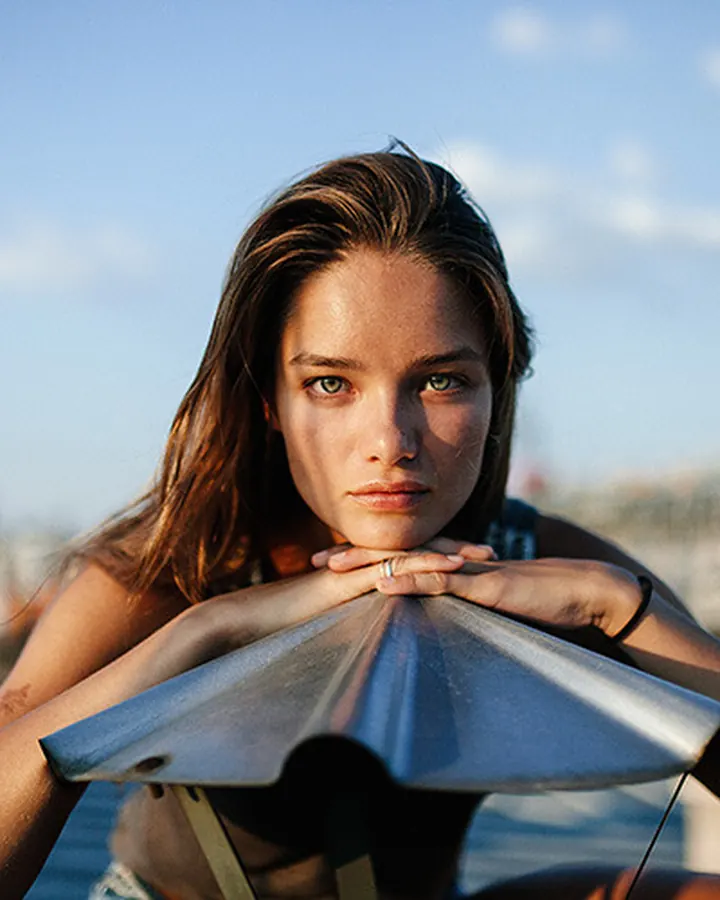 Jeune femme aux cheveux châtains et yeux bleus, posant avec les mains sous le menton devant un ciel bleu.