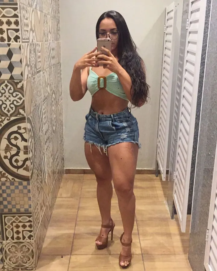 Femme aux longs cheveux noirs, portant un haut vert et un short en jean, prenant un selfie dans un couloir décoré de carreaux.