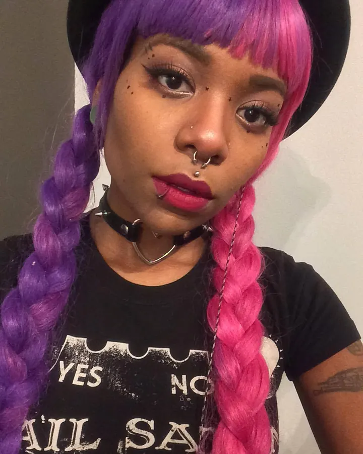 Femme trans aux cheveux châtains, vêtue d'une tenue sombre, assise sur un lit, regardant directement l'appareil.