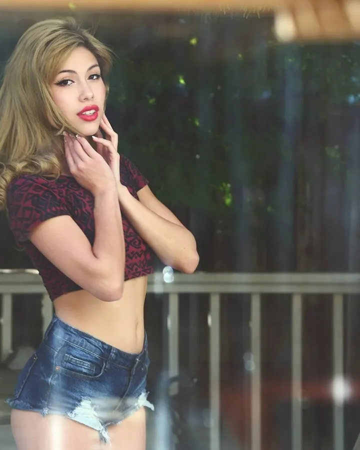 Jeune femme trans blonde, vêtue d'un haut rouge et d'un short en jean, posant devant une vitre avec une main sur son visage.
