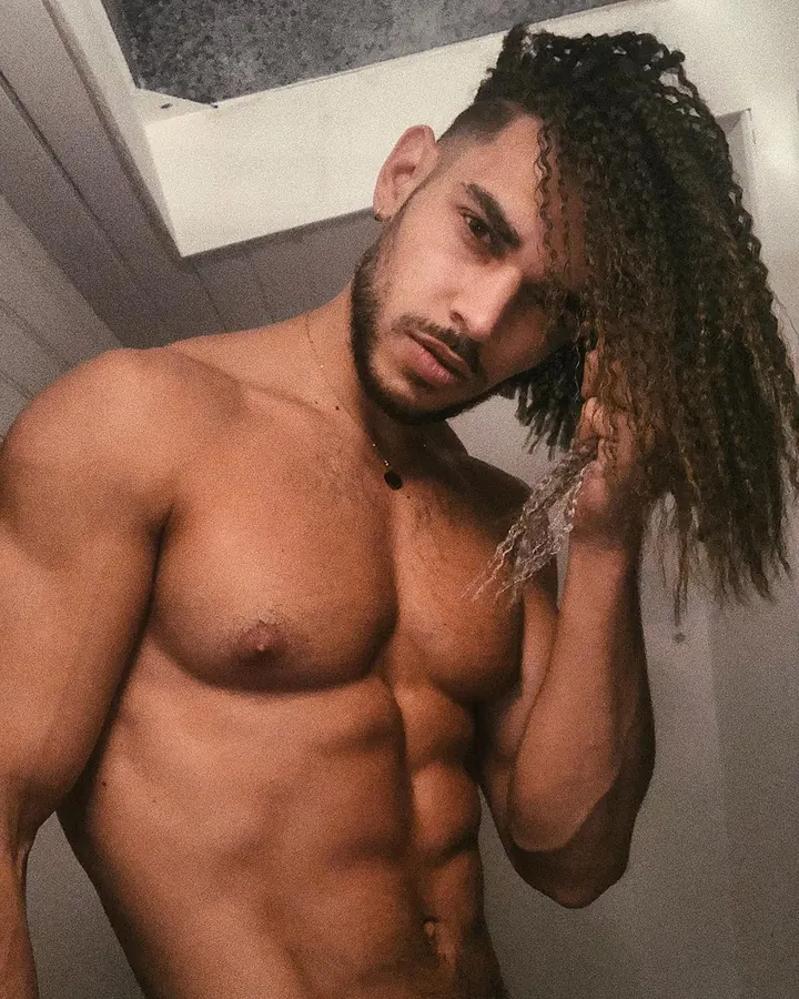 Femme trans aux cheveux châtains, vêtue d'une tenue sombre, assise sur un lit, regardant directement l'appareil.