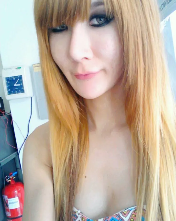 Femme trans blonde aux yeux bleus, vêtue d'une tenue sombre, assise sur un canapé, regardant l'appareil.