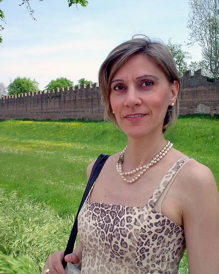 Femme aux cheveux bruns, portant un haut vert clair, tenant ses mains autour de son cou, souriant à la caméra.