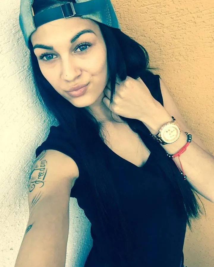 Femme aux cheveux noirs longs, portant une capuche, un haut noir, avec un tatouage 'Freedom' sur le bras, regardant l'appareil.