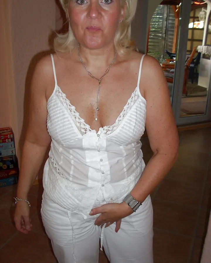 Femme blonde aux cheveux longs, portant une chemise verte à col dentelle, jeans, et plusieurs bagues, dans un cadre intérieur.