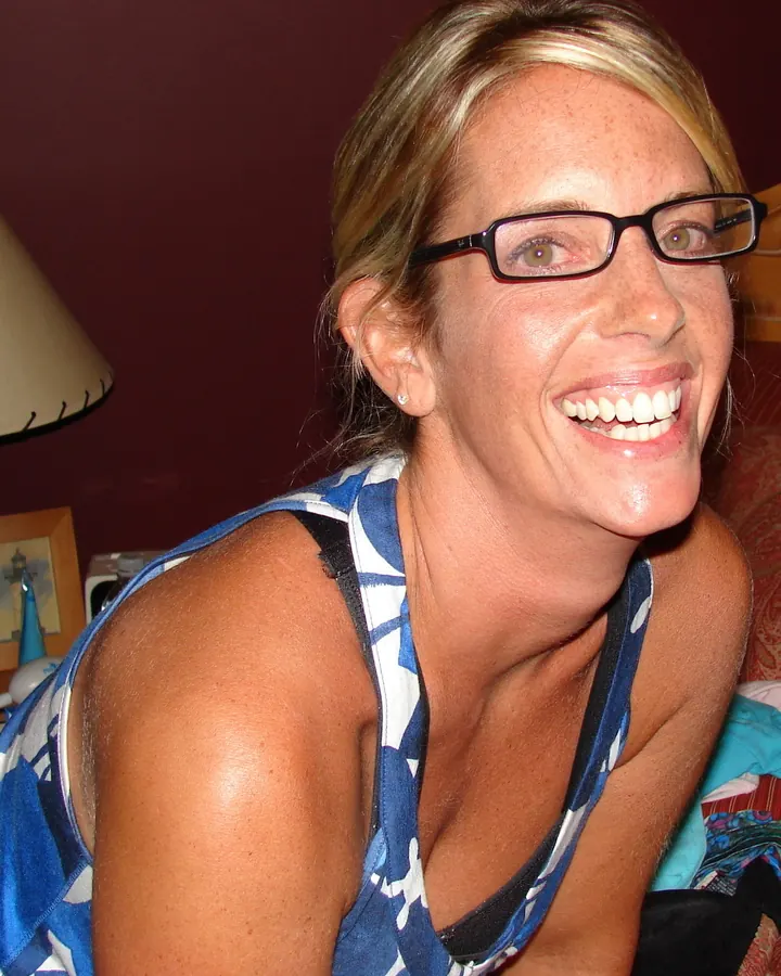 Femme blonde souriante, portant des lunettes à monture noire, un haut bleu et blanc à motifs, dans un cadre intérieur chaleureux.