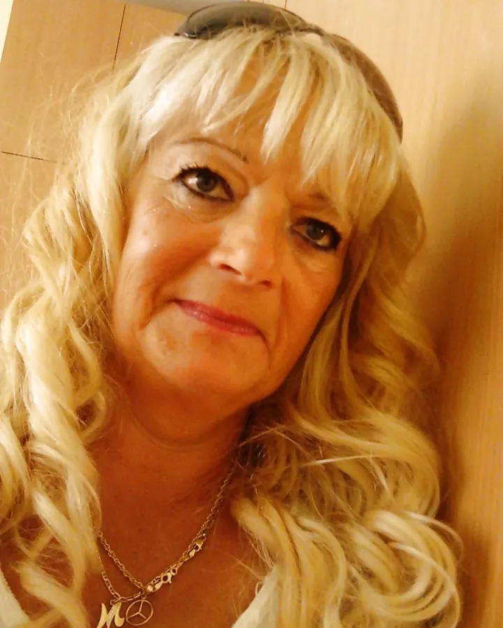 Femme aux cheveux blonds platine, yeux marrons, souriant légèrement, portant un collier avec un pendentif en forme de paix et un M.