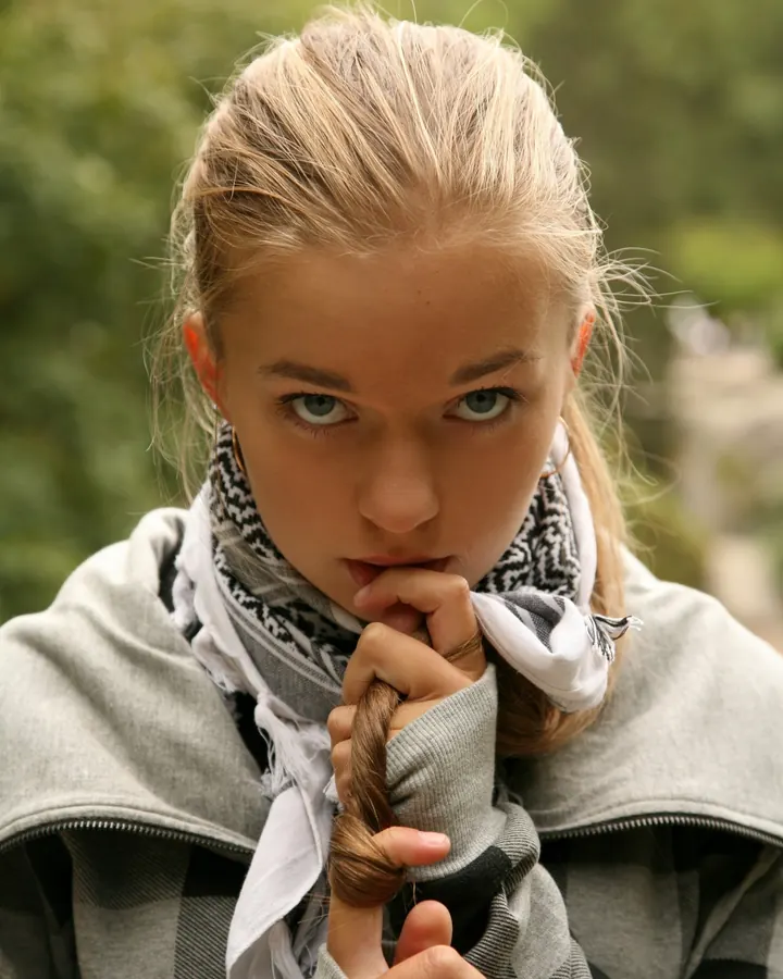 Jeune femme blonde aux yeux bleus, portant un sweat à capuche gris à carreaux et un foulard blanc et noir, tenant une mèche de cheveux dans sa bouche.