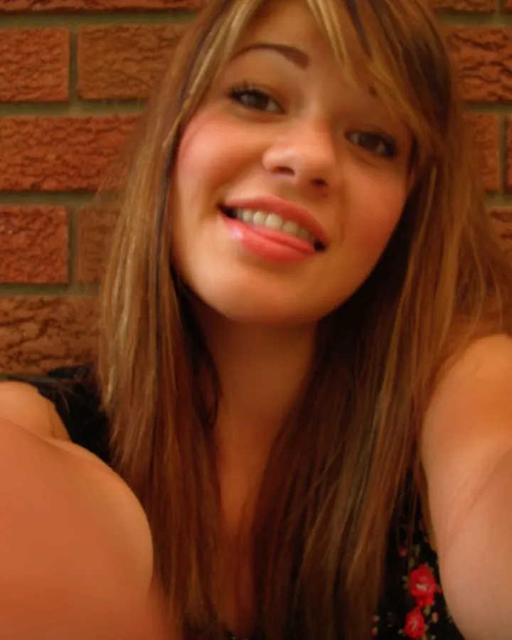 Jeune femme aux cheveux châtains prenant un selfie devant un mur de briques, souriante, vêtue d'un haut à fleurs.