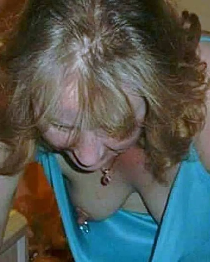 Femme aux cheveux blonds bouclés penchée en avant, portant un haut bleu, dans une pièce éclairée par une lampe.